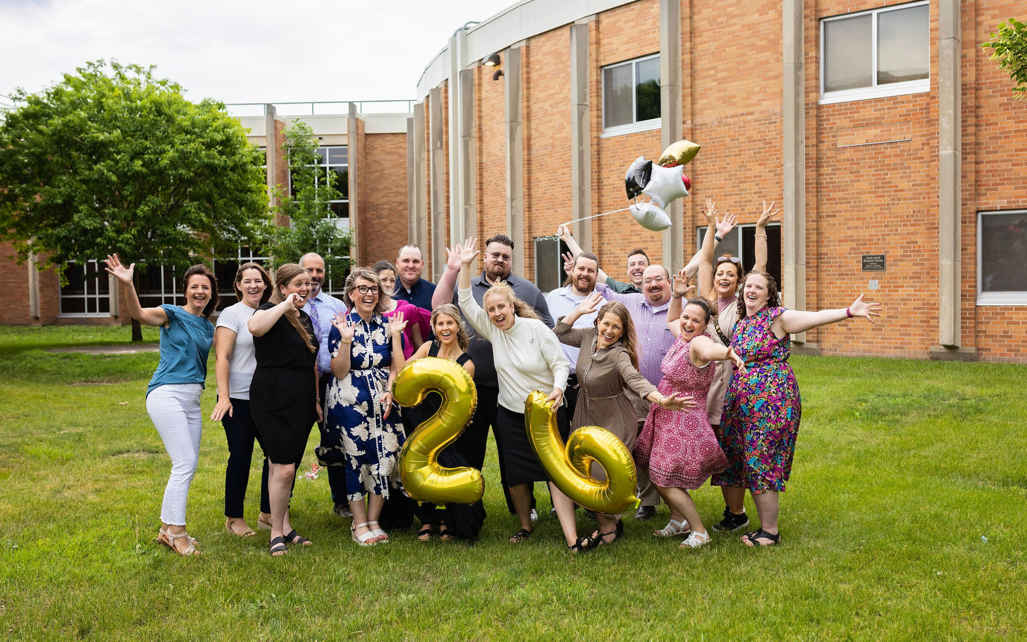 A joyful group photo of Falcon View Online school faculty celebrating on a lawn in front of a brick building. The central figure holds a large bouquet of gold, red, and silver graduation balloons.